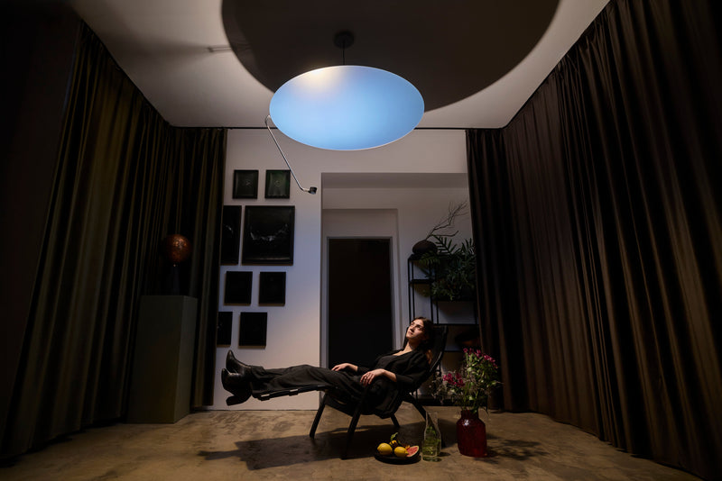 A woman reclines in a dark room under Sunday Light, with flowers and fruit in rich colour beside her