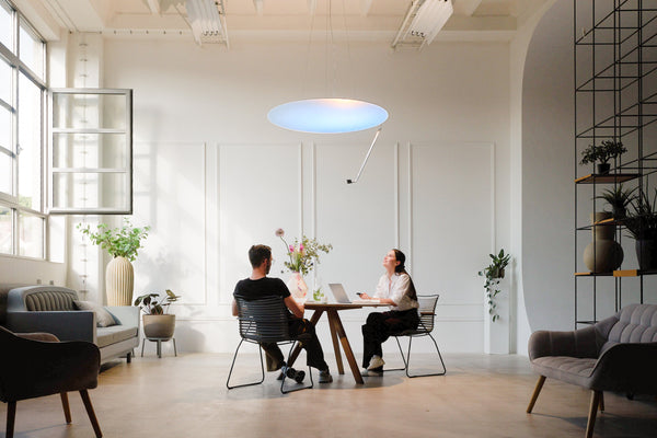 Two people at a table under Sunday Light in a white studio, one looking up at the light