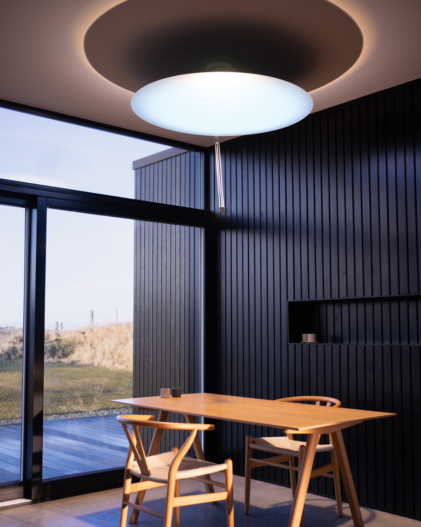 Sunday Light artificial skylight glowing with cool daylight above a wooden dining table in a black-panelled modern room
