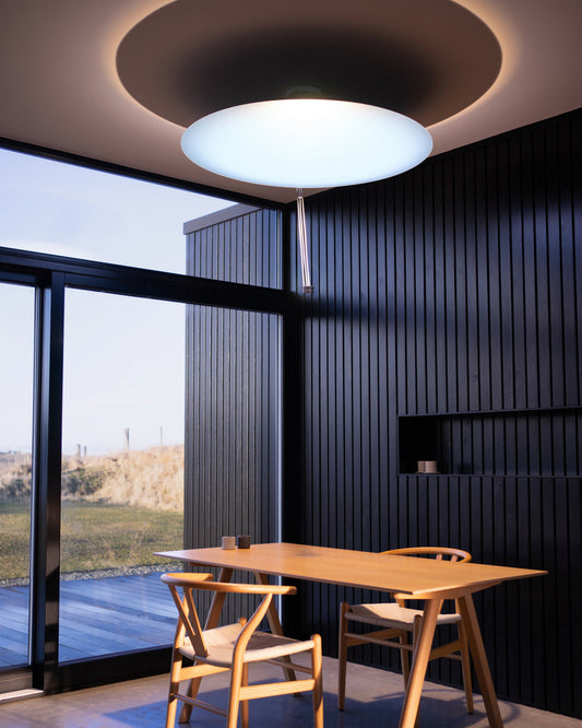 Sunday Light artificial skylight glowing with cool daylight above a wooden dining table in a black-panelled modern room